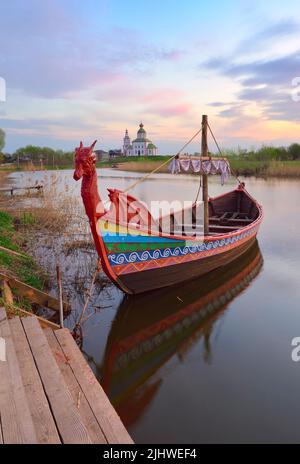 Boot auf dem Fluss Kamenka. Tourist drakkar in der Nähe des Ufers an der Iljinsky Kirche. Susdal, Russland, 2022 Stockfoto