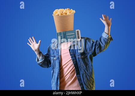 Glücklicher junger Mann mit einem Eimer knackigen Popcorns anstelle seines Kopfes und Kinokarte auf blauem Hintergrund Stockfoto