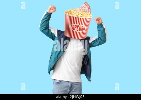 Glücklicher junger Mann mit einem Eimer knackigen Popcorns anstelle seines Kopfes und Kinokarten auf blauem Hintergrund Stockfoto
