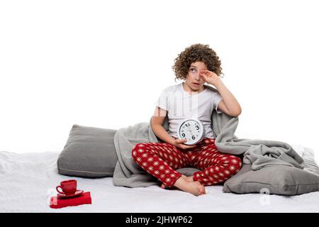Сurly kurzhaarige Mädchen in roten karierten Pyjamahosen und T-Shirt hält großen Wecker. Ein verschlenktes weibliches Kind, das sich im Studio mit weißem Hintergrund die Augen reibt Stockfoto