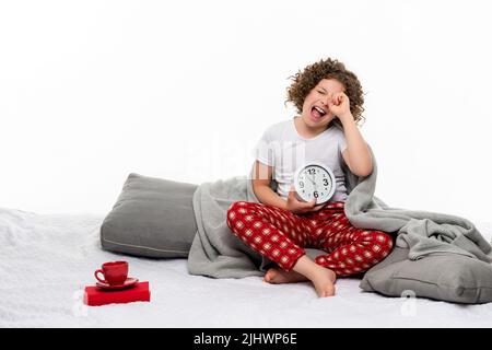 Сurly kurzhaarige Mädchen in roten karierten Schlafanzughosen und Basic-T-Shirt hält großen Wecker. Weibliches Kind gähnt auf dem Bett in weißem Hintergrund stu Stockfoto