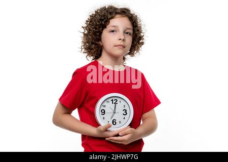 Сurly kurzhaarige Teenager-Mädchen trägt rote Basic-T-Shirt in weißen Hintergrund Studio. Verärgert weibliches Kind halten Wecker. Gesunder Schlaf und tägliche Routine. Stockfoto