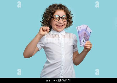 Unglaublich zufrieden präteen Mädchen in weißer Bluse und runde Brillen hält Stapel von 500 Euro-Banknoten auf hellblauen Studio-Hintergrund. Kinder Flosse Stockfoto