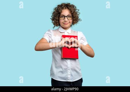 Mädchen mit lockigen Haaren 10-12s in weißer Bluse und runder Brille drückt sich rotes Lehrbuch an und macht Herzzeichen mit beiden Händen. Concep Stockfoto