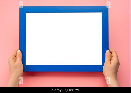 Nachbau des Haltrahmens. Foto-Mockup. Frau Hände halten blauen Rahmen. Für Rahmen- und Poster-Design. Rahmengröße A4. Isoliert auf rosafarbenem Hintergrund. Blanker Phot Stockfoto