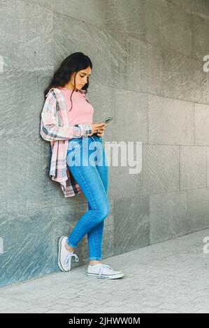 Trendy junge Teenager-Mädchen chatten auf ihrem Handy in den sozialen Medien, wie sie sich gegen eine graue Wand mit Copyspace lehnt steht Stockfoto