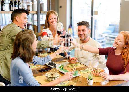 Eine Gruppe fröhlicher multirassischer Freunde, die sich gegenseitig angucken und mit Rotwein klirrende Weingläser anklirren, während sie gemeinsam am Tisch mit Essen feiern Stockfoto