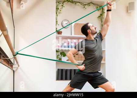 Seitenansicht eines sportlich starken Mannes im aktiven Ohr, der während des intensiven Trainings in einem leichten modernen Fitnessstudio mit Widerstandsbändern trainiert Stockfoto