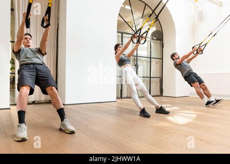 Vollkörper starker Menschen in der aktiven Trainingsbekleidung, die während des Funktionstrainings in einem modernen, leichten Fitnessstudio Stretching-Übungen mit TRX-Trägern machen Stockfoto