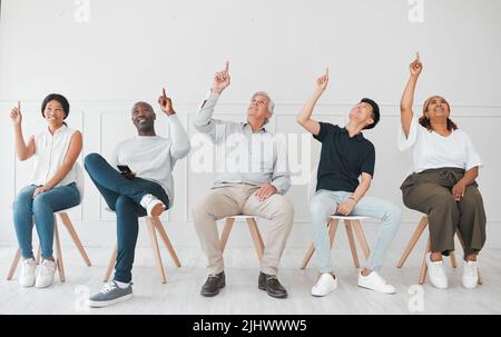 Jetzt ist das faszinierend. Eine vielfältige Gruppe von Menschen, die nach oben zeigen, während sie in einer Reihe vor einem weißen Hintergrund sitzen. Stockfoto