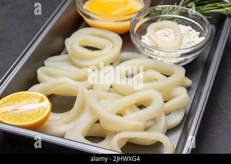 Tintenfisch in Ringe auf der Palette geschnitten. Geschlagene rohe Eier, Butter und Mehl in Schüsseln. Draufsicht. Schwarzer Hintergrund Stockfoto