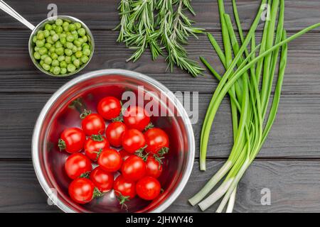 Tomaten und frische grüne Erbsen in Metallschüsseln. Grüne Zwiebeln und Rosmarin. Flach liegend. Dunkler Holzhintergrund Stockfoto