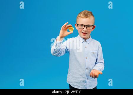 Kryptowährung und moderne Finanzwirtschaft. Fröhlicher Junge in Brillen mit goldenem Bitcoin im Studio auf blauem Hintergrund. Zukünftiger Geschäftsmann Stockfoto