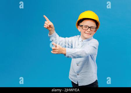 Netter Junge Bauarbeiter in gelbem Hut, der im Innenstudio auf blauem Hintergrund steht und mit den Fingern in Richtung zeigt. Zukünftige Baumaßnahmen Stockfoto