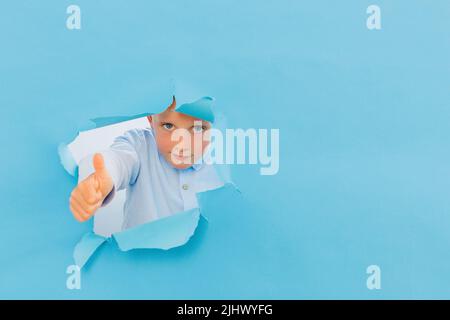 Happy cute boy hat Spaß auf blauem Hintergrund Wand gespielt, klettert durch ein Loch in das Papier. Helle und lustige Emotionen des Jungen Stockfoto