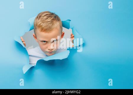 Happy cute boy hat Spaß auf blauem Hintergrund Wand gespielt, klettert durch ein Loch in das Papier. Helle und lustige Emotionen des Jungen Stockfoto