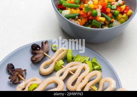 Tintenfischringe, Babykrake auf Holzspießen auf grauem Teller. Gemüse in Schüssel mischen. Draufsicht. Weißer Hintergrund. Stockfoto