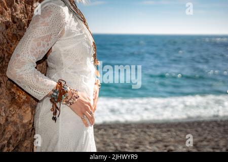 Frau in böhmischer Kleidung am Strand bei Sonnenuntergang. Boho-Stil für modischen Look im Resort. Frau mittleren Alters gut aussehende Frau in weißem Kleid und Boho-Stil Stockfoto