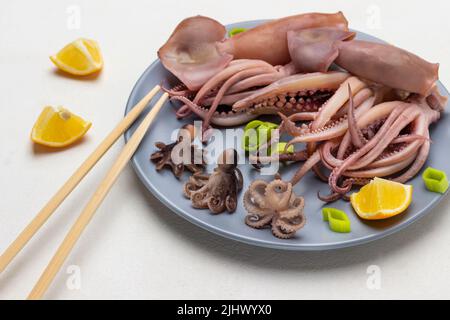 Gekochte Babykrake und Tintenfisch mit Tentakeln auf grauem Teller. Bambusstäbchen auf dem Teller. Zitronenscheiben auf dem Tisch. Draufsicht. Weißer Hintergrund Stockfoto