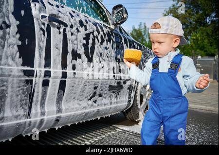 Nahaufnahme eines Kleinkindes mit ernsthaftem Gesichtsausdruck, das neben einem schwarzen, schaumbedeckten Auto steht und das Auto verwirrt anschaut. Kleiner Junge, der neue Lektion lernt. Stockfoto