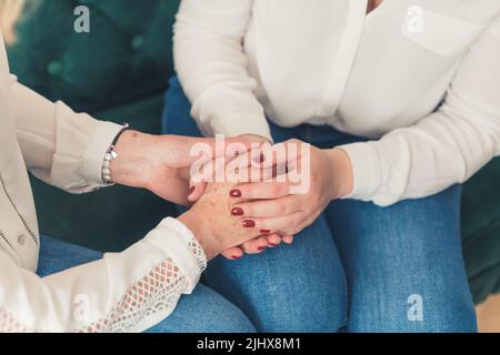 Zwei nicht erkennbare weiße Frauen sitzen auf der Couch und halten sich die Hände. Weißes Hemd und Jeans. Rote Nägel. Mutter und Tochter. Hochwertige Fotos Stockfoto