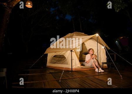 Attraktive und schöne junge asiatische Frau, die vor ihrem Zelt sitzt, heißen Kaffee schlürft und den schönen Nachthimmel anschaut. Camping c Stockfoto