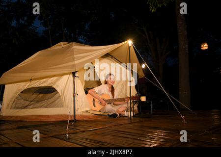 Attraktive junge Asiatin, die nachts vor dem Tipi-Zelt auf dem Campingplatz Gitarre spielt. Outdoor-Aktivitätskonzept. Stockfoto
