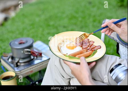 Camper-Reisende, die ihr Frühstück isst, während sie auf dem Campingplatz campen. Nahaufnahme eines Frühstückssets in den Händen der Frau Stockfoto