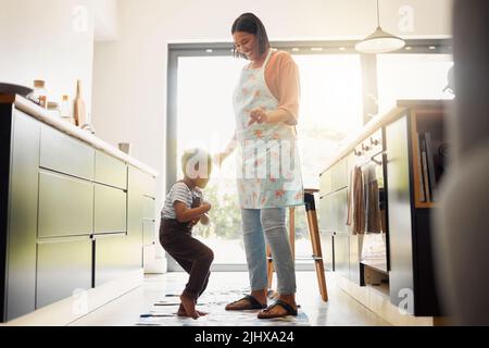 Ein kleiner gemischter Rennjunge, der tanzt und Spaß mit seiner Mutter in einer Küche hat. Glückliche Familie von zwei Kindern, die beim Backen in einer Küche Begeisterung ausdrückt Stockfoto