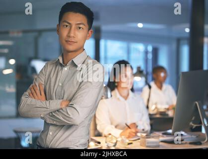 Ein junger Geschäftsmann, der spät in der Nacht mit gekreuzten Armen in einem modernen Büro steht. Stockfoto