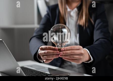 Geschäftsfrau Hält Glühbirne Mit Beiden Händen Im Büro. Frau Im Anzug, Die Licht Zwischen Den Palmen Auf Dem Schreibtisch Mit Lap Top Hat Und Wichtig Präsentiert Stockfoto