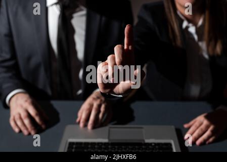 Sitzender Mann Auf Dem Schreibtisch Mit Lap Top Und Frau, Die Mit Einem Finger Auf Wichtige Botschaften Zeigt. Geschäftsfrau Präsentiert Krutialinformationen Mit Einer Hand Stockfoto