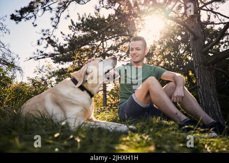 Mann mit seinem Hund im Gras unter dem Baum sitzend. Tierbesitzer streichelte seinen labrador Retriever an sonnigen Sommertagen. Stockfoto