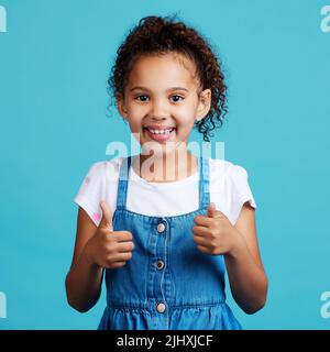 Weiter so. Ein entzückendes kleines Mädchen zeigt Daumen nach oben, während sie vor einem blauen Hintergrund steht. Stockfoto