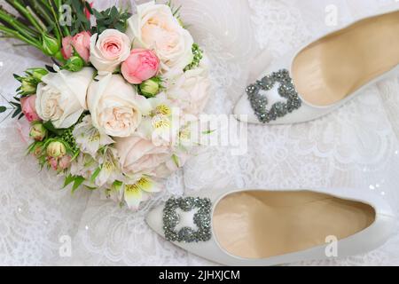 Hochzeitskomposition mit weißen Seidenschuhen, pastellfarbenem Brautstrauß auf weißem, verschwommenem Hintergrund. Draufsicht Stockfoto