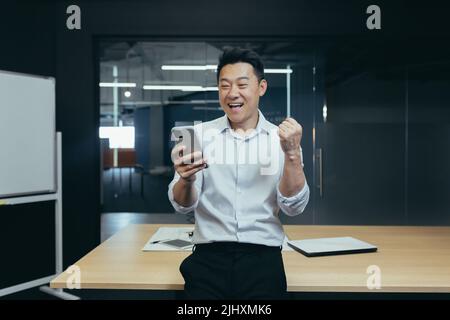Erfolgreicher Geschäftsmann aus dem Finanzwesen, Asian, der im Büro steht, gute Nachrichten vom Telefon liest, die Hand hochhält und Sieg und Triumph feiert Stockfoto