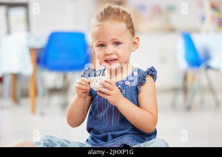 Portrat eines entzückenden kleinen kaukasischen Mädchens, das allein in der Vorschule sitzt und eine kleine Teetasche hält. Lächelndes Kind beim Spielen und einer Teeparty Stockfoto