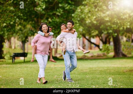 Gemischte Rennfamilie im Park. Glückliche junge Mutter und Vater huckepack ihren Sohn und ihre Tochter draußen. Das Paar trägt seine Kinder während des Spazierens Stockfoto