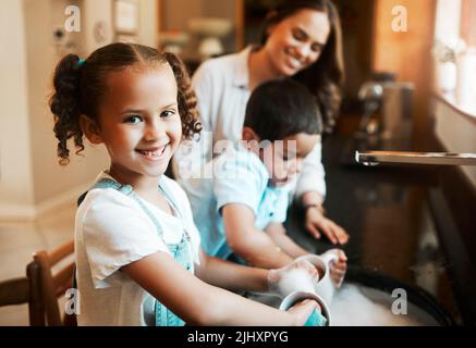 Fröhliches kleines gemischtes Rennmädchen, das ihrer Mutter hilft, das Geschirr in der Küche zu Hause zu waschen. Hispanische Kinder lächeln beim Waschen einer Tasse mit Seife Stockfoto