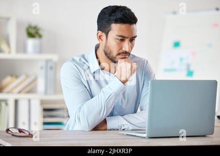 Junger seriöser Geschäftsmann mit gemischter Rasse, der an einem Tisch sitzt und allein in einem Büro auf einem Laptop arbeitet. Ein hispanischer männlicher Chef liest und sendet Stockfoto