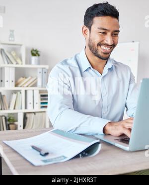 Junger glücklicher Geschäftsmann mit gemischtem Rennen, der allein in einem Büro auf der Arbeit an einem Laptop arbeitet. Ein hispanischer männlicher Chef lächelt, während er eine E-Mail tippt und eine verwendet Stockfoto