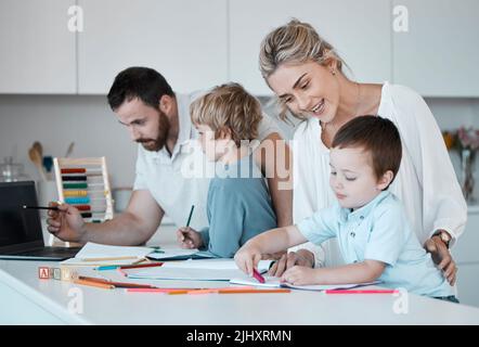 Die junge kaukasische Familie hat ihre beiden Kinder zuhause. Liebevolle Eltern helfen ihren kleinen Kindern bei den Hausaufgaben. Mama und Papa lehren Kinder Stockfoto