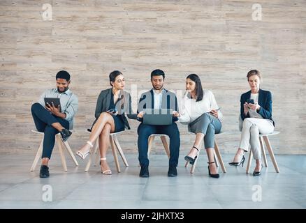 Unser Unternehmen kann nicht ohne Technologie laufen. In voller Länge eine Gruppe von Geschäftsleuten, die in einer Reihe sitzen und Technologie einsetzen. Stockfoto