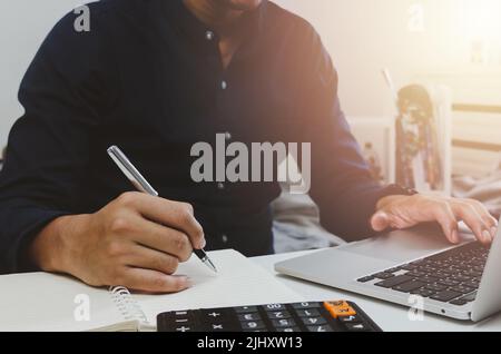 Geschäftsmann Hand hält Stift Schreibbuch und Tastatur Computer Laptop auf dem Schreibtisch. Stockfoto