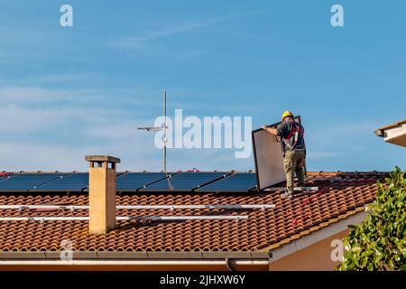 Ein Techniker hebt eine Solaranlage an, um eine Photovoltaikanlage auf einem rot gefliesten Dach zu installieren Stockfoto