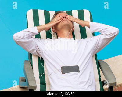 Ein weißer Mann in einem weißen Hemd liegt mit den Händen über den Augen auf einer Couch, während er mit Kopfhörern von einem Smartphone Musik hört Stockfoto