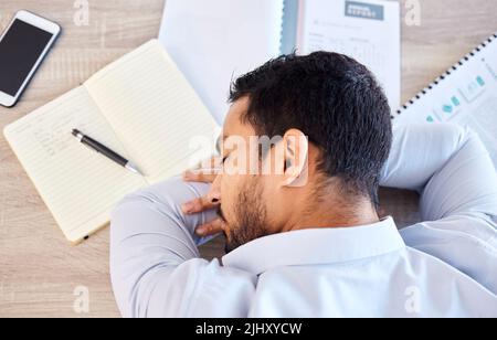 Geschäftsmann schläft von oben auf einem Tisch. Ein müder junger Mann, der bei der Arbeit in einem modernen Büro ein Nickerchen auf seinem Schreibtisch macht. Draufsicht auf einen erschöpften Mitarbeiter Stockfoto