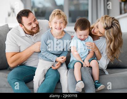 Sorglose, liebevolle Eltern kitzeln und verbinden sich mit niedlichen kleinen lachenden Söhnen. Lächelnde kaukasische vierköpfige Familie, die sich zu Hause auf dem Sofa entspannt.Verspielt Stockfoto