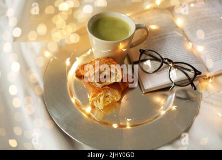 Croissants, Matcha-Tee, Buch und Gläser im Bett Stockfoto