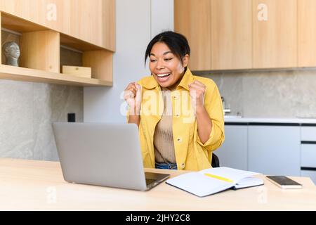 Überglücklich junge Frau überrascht Sieg schaut auf den Laptop im Büro sitzen, weiblich feiert gute Nachrichten per E-Mail erhalten, bekam ein Stellenangebot, erstaunt Frau jubeln gewinnen in einer Online-Lotterie Stockfoto